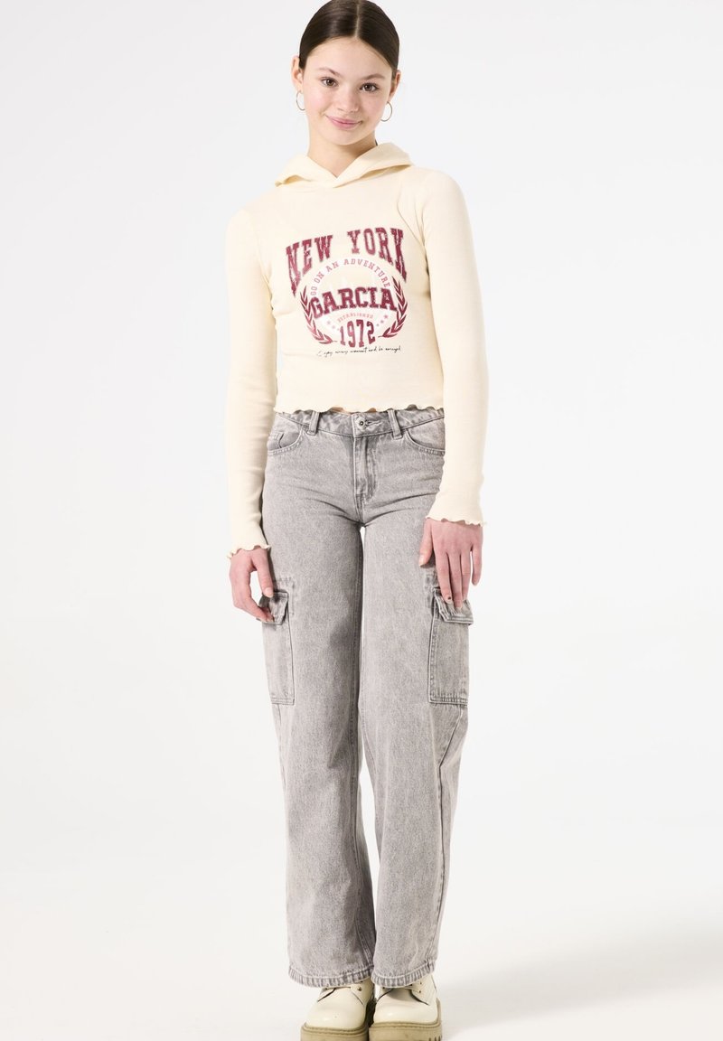 Sudadera con capucha crema y texto gráfico rojo, combinada con pantalones cargo grises de pierna ancha con bolsillos laterales. El modelo lleva zapatos beige.