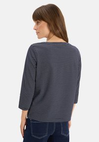 Femme aux cheveux bruns portant un haut à rayures bleu marine et blanches à manches longues et un jean bleu foncé, tournée légèrement vers la gauche devant un fond uni.