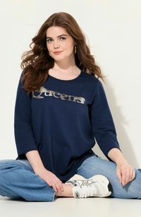 Chemise à manches longues marine avec le texte argenté "Queens", associée à un jean bleu clair ample et des baskets blanches avec des accents argentés.
