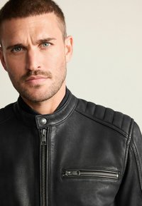 Zwarte leren jas met een hoge kraag, ritssluiting aan de voorkant en geribbelde accenten op de schouders. Voorzien van een borstzak met ritssluiting. Glad van textuur.