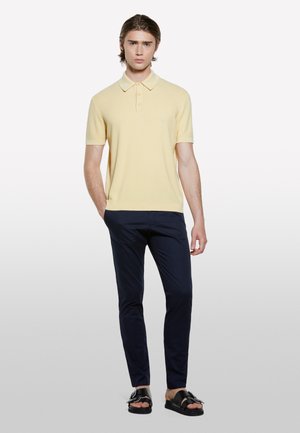 Polo shirt jaune à manches courtes en maille texturée, associé à un pantalon slim-fit bleu marine et des sandales noires à boucles.