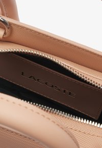 Die Innenseite der Lacoste-Handtasche weist ein glattes, sandfarbenes Lederetikett auf. Das Material der Tasche ist strukturiert mit einem dezenten Muster und einem silbernen Reißverschluss.