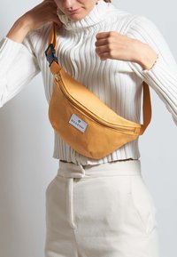 Marsupio giallo senape realizzato in tessuto resistente, dotato di una tasca con cerniera e una patch con logo. Viene indossato a spalla con una tracolla regolabile.