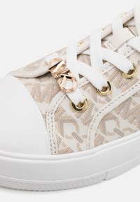 Beige kuviollinen lenkkari, jossa on valkoinen kuminen kärki, valkoiset nauhat, kultaiset rei'itetyt osat ja koriste, jossa on "MK" kultaisena.