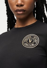 Κλείσιμο εικόνας ενός ατόμου που φοράει μαύρο μπλουζάκι με στρογγυλή μπεζ λογότυπο Versace Jeans Couture στο αριστερό στήθος.