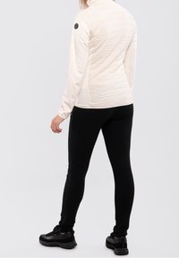 Personne portant une veste longue à manches longues texturée crème, un pantalon slim noir et des baskets noires, se tenant de dos contre un fond uni.