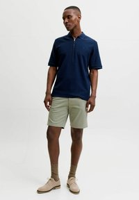 Άνδρας με μπλε σκούρο κοντομάνικο πόλο, μπεζ σορτς, καφέ κάλτσες και ταμπα παπούτσια, στέκεται μπροστά σε απλό λευκό φόντο.