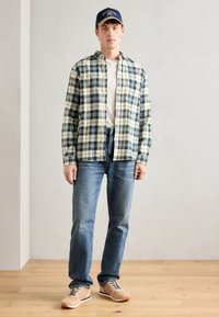 Chemise à carreaux verte et beige, t-shirt blanc en dessous, jeans bleus et chaussures beiges avec des accents noirs. Les accessoires comprennent une casquette bleue.