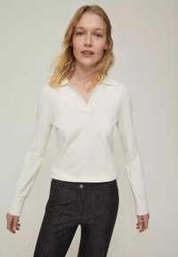 Maglione bianco a maniche lunghe con scollo a V e colletto, abbinato a pantaloni slim-fit scuri e strutturati. Il materiale sembra morbido con polsini a coste.