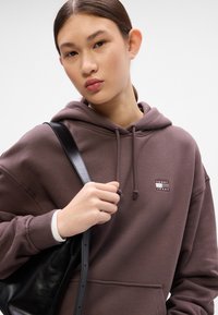 Hnědá mikina s kapucí a šňůrkou, s žebrovanými manžetami a přední kapsou. Obsahuje malé grafické logo. Model drží černou kabelku přes rameno.
