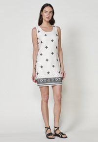 Weißes ärmelloses Kleid mit schwarzen geometrischen Mustern, das ein strukturiertes Saumdesign aufweist. Getragen mit schwarzen Riemchensandalen.