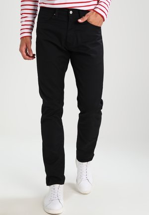 Person iført sorte bukser, hvide sneakers og en rød-hvid stribet langærmet trøje, med den ene hånd i lommen og går fremad.
