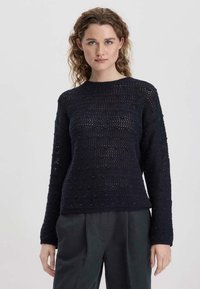 Marineblauer Strickpullover mit strukturiertem Design, featuring offene Maschen und erhabene Punkte. Klassischer Rundhalsausschnitt und gerippte Säume, über dunklen Hosen getragen.
