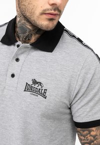 Hallhalli hallitav polo särk, millel on must krae ja mansetid, kolm nuppu ning Lonsdale logo. Omab musti külgmisi aktsente, millel on brändimine õlal.