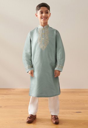 Jongen glimlachend en staand, draagt een lichtblauwe traditionele geborduurde kurta met witte broek en bruine schoenen op een houten vloer.