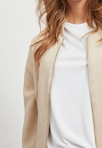 Frau mit einer beigen Zip-Jacke über einem weißen Rundhals-Shirt, sichtbare wellige, hellbraune Haare, die von den Schultern bis zum Nacken reichen.