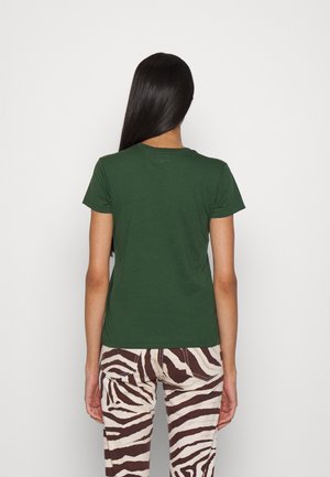 Frau mit langen schwarzen Haaren steht vor einer neutralen Wand und trägt ein schlichtes dunkelgrünes T-Shirt mit kurzen Ärmeln sowie Hose mit Zebramuster.