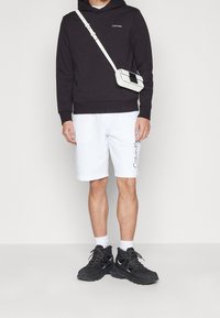 Zwarte hoodie, witte short met trekkoorden en zijlogo, en zwarte sportschoenen. Inclusief een kleine witte crossbody tas.
