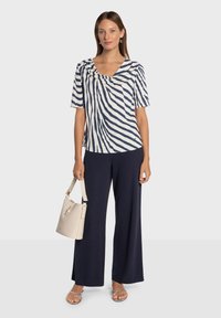 Granatowa i biała bluzka w paski z dekoltem typu drapowany, zestawiona z granatowymi spodniami o szerokich nogawkach. Modelka trzyma beżową torebkę na ramię i nosi srebrne sandały.