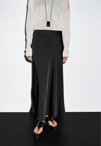 Frau trägt einen weißen Pullover, einen langen schwarzen Satinsrock, schwarze Plateausandalen und eine schwarze Anhängerkette, steht vor einem weißen Hintergrund.