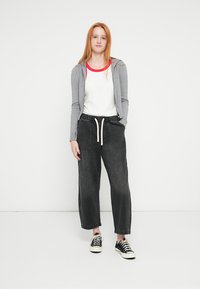 Γκρι φούτερ με φερμουάρ, λευκό πουκάμισο με κόκκινες λεπτομέρειες, μαύρο παντελόνι ευρύ στη βάση με ελαστική μέση και μαύρα αθλητικά παπούτσια. Τοποθετημένο πάνω σε απλό φόντο.