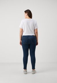 Mujer de pie de espaldas, vistiendo una camiseta blanca, jeans ajustados de un azul oscuro y zapatillas deportivas blancas, contra un fondo liso.