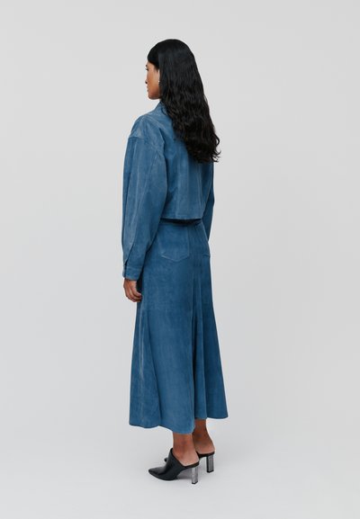 Completo in suede blu che include una giacca corta e una gonna fluida lunga fino alla caviglia. Le mule con tacco a blocco completano il look.