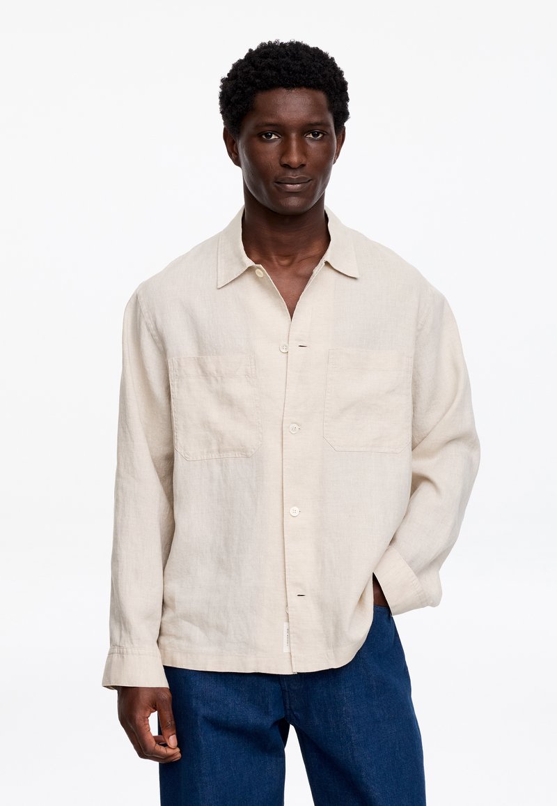 Homme à la peau foncée avec cheveux courts et bouclés portant une chemise en lin beige boutonnée et un jean bleu sur un fond blanc uni.