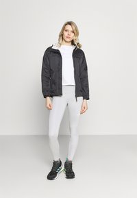 Chaqueta impermeable negra con capucha, llevada sobre una camiseta cortada blanca y leggings gris claro. Botas de senderismo negras con acentos verdes.