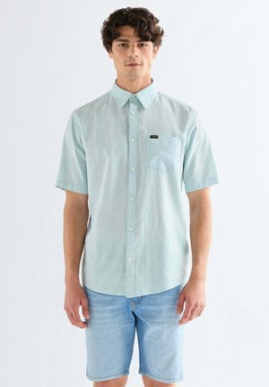 Młody mężczyzna stoi, ubrany w jasnoniebieską, krótkorękawową koszulę na guziki oraz jasne denimowe szorty na jednolitym tle.