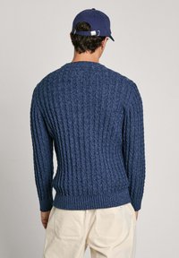 Marineblaue gerippte Strickjacke mit Rundhalsausschnitt und langen Ärmeln, die ein strukturiertes Muster und eng anliegende Bündchen aufweist. Getragen über hellfarbigen Hosen.