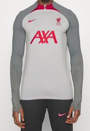 Man met een grijze en rode trainingsshirt van Liverpool FC met AXA sponsornaam en Nike-logo, gecombineerd met een zwarte broek met een roze Nike-logo.