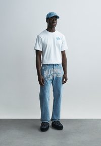 Λευκό μπλουζάκι με μπλε λογότυπο, τζιν παντελόνι με το λευκό γραφικό "Billionaire Boys Club" και μπλε καπέλο· συνδυασμένα με μαύρα παπούτσια με σχέδιο.