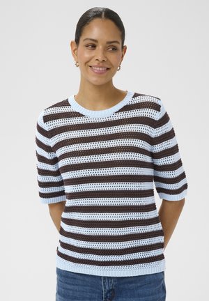 Mujer sonriente con el cabello oscuro recogido, lleva pendientes de aro, un top de punto de manga corta con rayas azules y marrones y vaqueros azules.