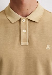 Beżowa koszulka polo wykonana z fakturowanej tkaniny, z pointed kołnierzykiem, zapięciem na trzy guziki oraz małym haftowanym logo na klatce piersiowej.