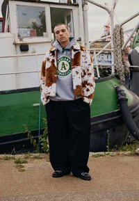 Νεαρός άνδρας με καφέ και λευκή γούνα, γκρι φούτερ Oakland Athletics, μαύρο φαρδύ παντελόνι και μαύρα παπούτσια, ο οποίος στέκεται κοντά σε ένα πράσινο και λευκό σκάφος.