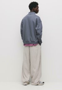 Felpa grigia oversize con colletto alto sovrapposta a una camicia viola, abbinata a pantaloni larghi di un beige chiaro e sneakers.