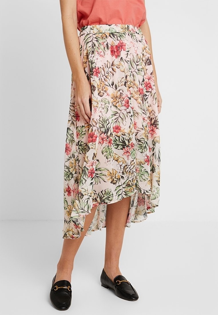 Femme portant une jupe fluide asymétrique à fleurs avec des feuilles roses, vertes et jaunes, associée à des mocassins noirs et un haut corail.