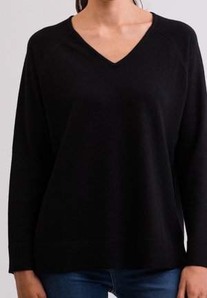 Kvinde iført en almindelig sort langærmet V-hals sweater og blå jeans står mod en lys baggrund.