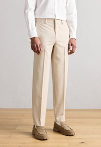 Pantalones beige con un corte recto, confeccionados en una tela suave. Cuentan con bolsillos laterales y un diseño frontal liso. Combinados con zapatos marrones.