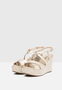 Sandali wedge metallici dorati con cinturini incrociati e fibbia regolabile alla caviglia. Texture intrecciata sul tacco wedge e design aperto arrotondato.