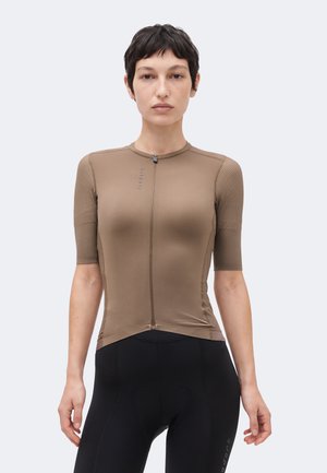 Mujer con pelo corto y oscuro, vistiendo un maillot de ciclista marrón ajustado de manga corta y pantalones cortos de ciclismo negros, de pie frente a un fondo liso.