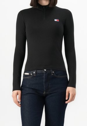 Kobieta ubrana w dopasowany czarny top z długim rękawem z małym logo Tommy Jeans oraz ciemnoniebieskie jeansy z wysokim stanem.