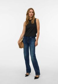 Mustast tekstuurse viimistlusega varrukateta top, kombineeritud tumesiniste lehvikpükstega ja mustade teravate kontsakingadega, käes pruun käekott.