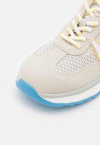 Baskets gris clair avec une tige en daim, des panneaux en maille, des lacets jaunes et blancs, et une semelle en caoutchouc bleu pour l'adhérence.