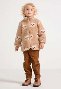 Faux fur beige jacket with cat pattern, zip closure, and side pockets. Paired with brown corduroy pants and brown lace-up boots.