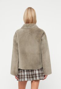 Manteau en fausse fourrure gris clair, avec un large col et des manches amples, montré de dos sur une jupe à carreaux marron. Texture douce.