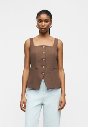 Kvinde med flettet hår iført en brun, ærmeløs, knappet top og lyseblå jeans mod en ensfarvet baggrund.