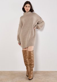 Beige übergroßes, gestricktes Kleid mit hohem Kragen, gerippten Bündchen und Saum. Kombiniert mit kniehohen, glatten braunen Lederstiefeln.