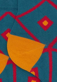 Calzini di cotone con una base teal, decorati con motivi a rombi rossi e dettagli arancioni sul tallone. Texture liscia con un design geometrico colorato.